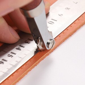 May include: A person using a leather stitching tool to create a line of stitching on a piece of brown leather. The tool has a metal wheel with teeth and a handle. The leather is being held in place with a ruler.