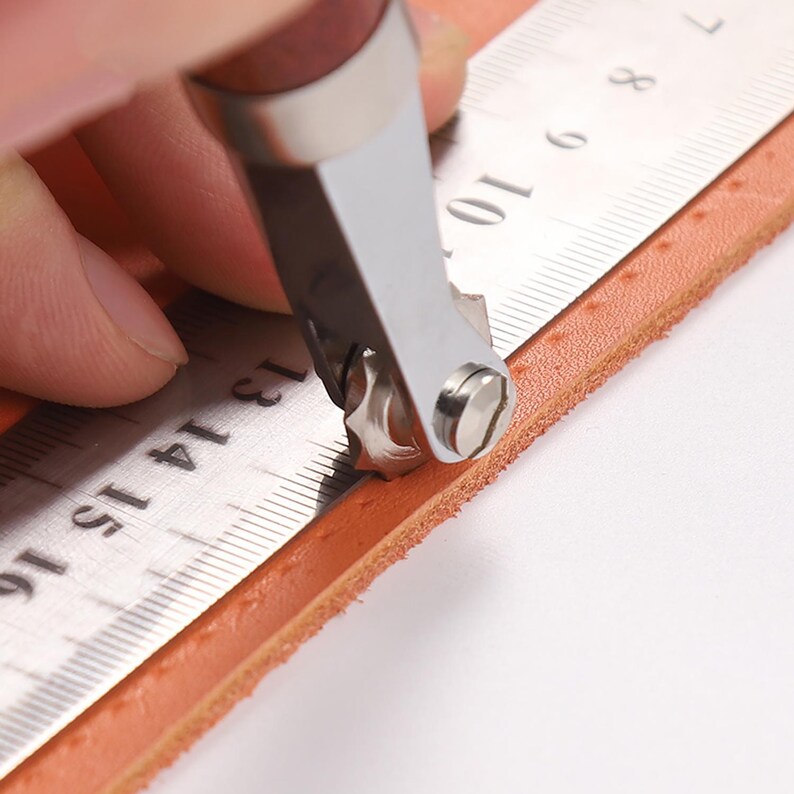 May include: A person using a leather stitching tool to create a line of stitching on a piece of brown leather. The tool has a metal wheel with teeth and a handle. The leather is being held in place with a ruler.