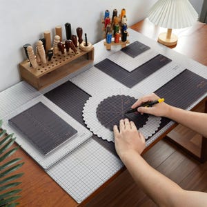 May include: A collection of gray and black grid cutting mats in various shapes, including rectangular and circular designs, arranged on a wooden table. A wooden tool organizer holds several leatherworking tools, and a rack displays colorful thread spools.