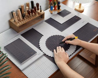 May include: A collection of gray and black grid cutting mats in various shapes, including rectangular and circular designs, arranged on a wooden table. A wooden tool organizer holds several leatherworking tools, and a rack displays colorful thread spools.
