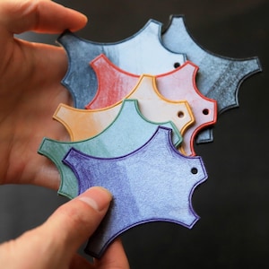 May include: A selection of multi-coloured, abstract-shaped tags in shades of blue, red, orange, green, and purple. Each tag features a small hole and a distinctive, textured finish. The tags are held in a hand against a dark backdrop.