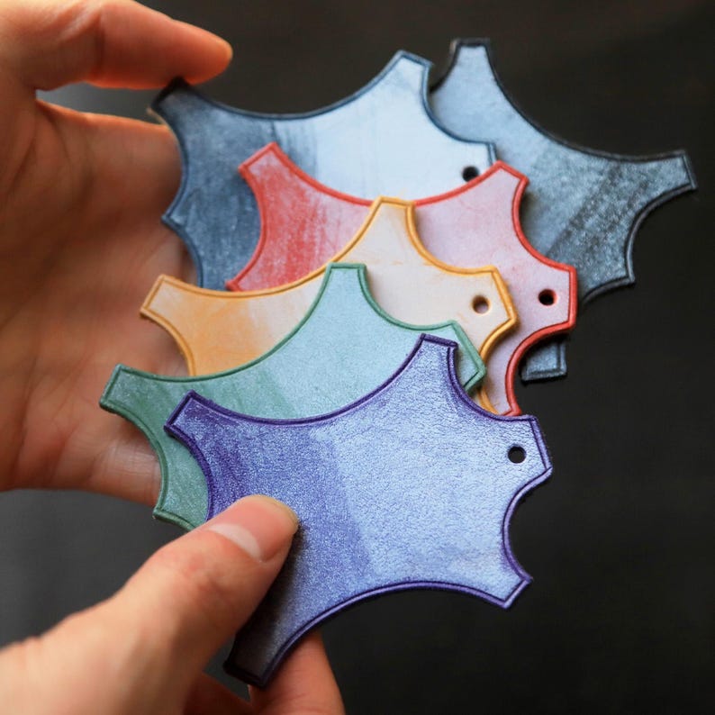 May include: A selection of multi-coloured, abstract-shaped tags in shades of blue, red, orange, green, and purple. Each tag features a small hole and a distinctive, textured finish. The tags are held in a hand against a dark backdrop.