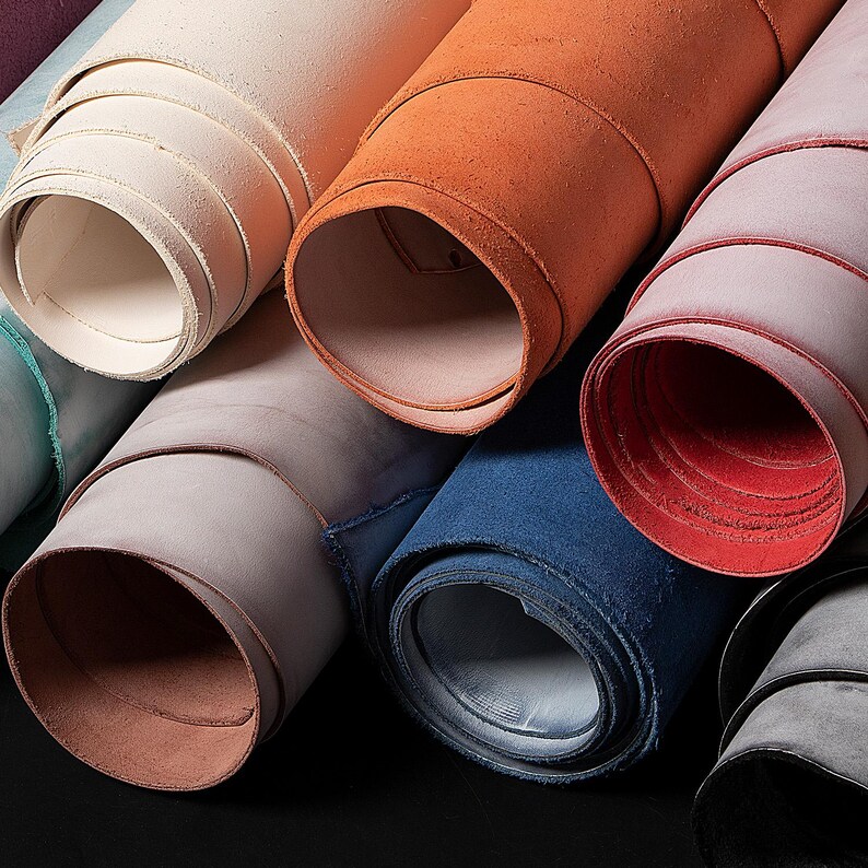 May include: A selection of rolled leather in various colours. The rolls are in shades of cream, orange, red, blue, and grey. The leather has a textured appearance, possibly suede, with visible grain. The rolls are tightly wound, showing the edges of the material.