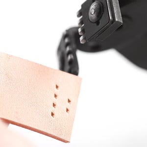 May include: Close-up of a leather crafting tool punching holes in a piece of light brown leather. The tool is black and metallic, with a series of sharp edges. The leather has a textured surface and the holes are evenly spaced.
