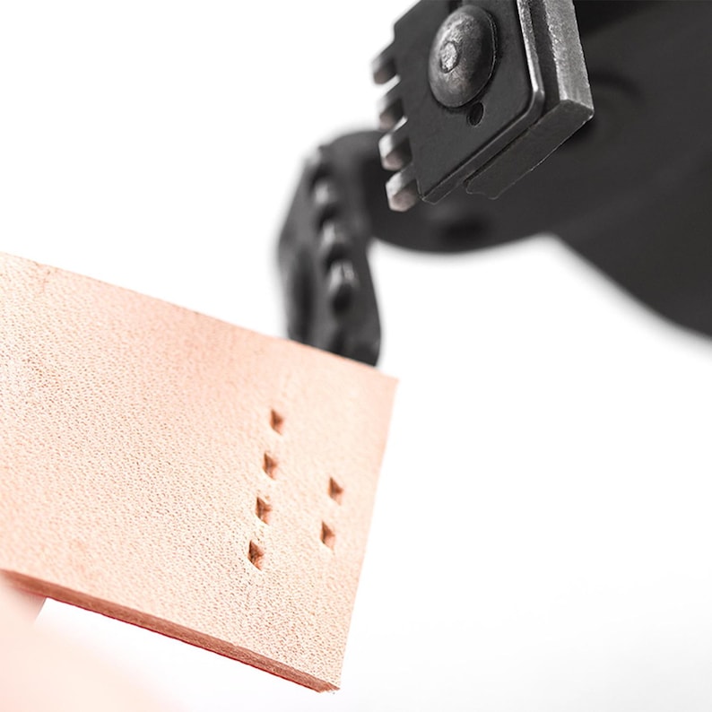May include: Close-up of a leather crafting tool punching holes in a piece of light brown leather. The tool is black and metallic, with a series of sharp edges. The leather has a textured surface and the holes are evenly spaced.