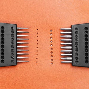 May include: Two black leather stitching tools with rows of needles on a brown leather surface. The tools are used to create evenly spaced holes in leather for stitching.
