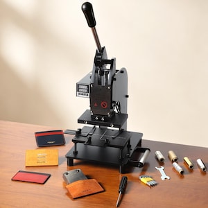 May include: A black leather embossing machine with a digital display, handle, and various tools. Several leather wallets in red, blue, and tan are arranged on a wooden surface, showcasing the machine's capabilities.