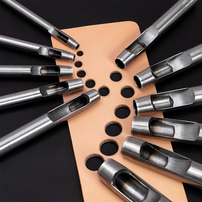 May include: A set of ten silver leather hole punches of varying sizes, arranged in a fan shape on a piece of tan leather with a series of punched holes.