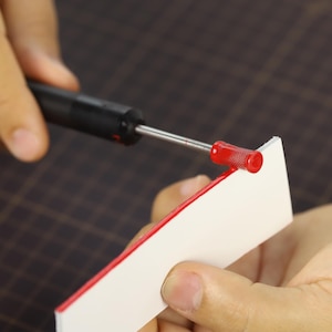 May include: A person using a red and silver tool to apply red paint to a white surface.