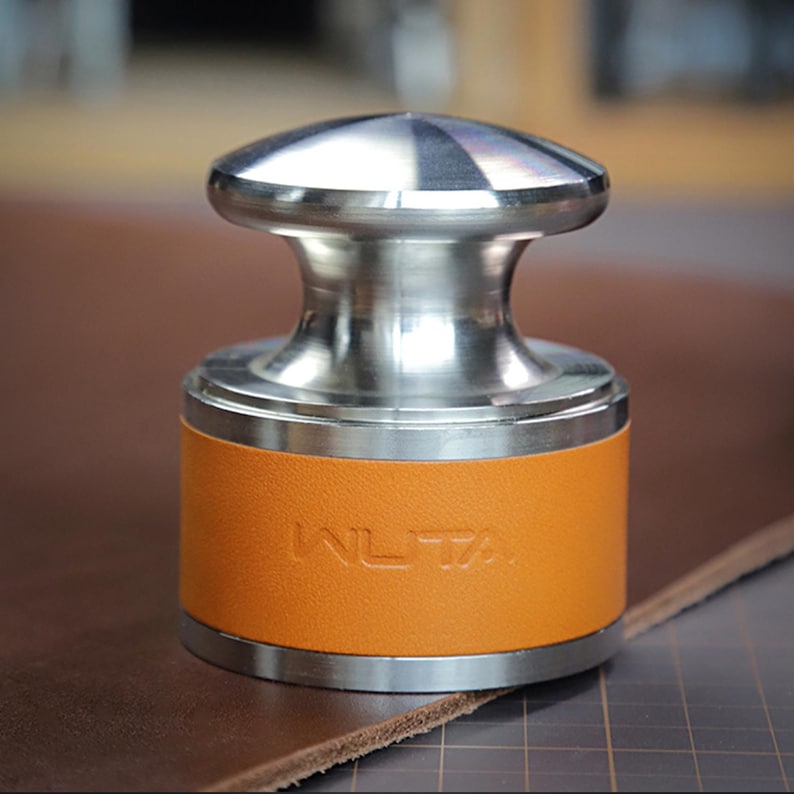 May include: A metal leathercraft tool with a stainless steel top and base, and an orange leather-wrapped body. The word "WLTA" is embossed on the leather. The tool is designed for leatherworking.
