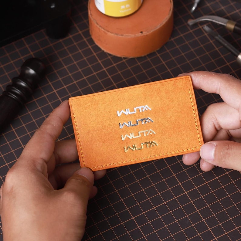 May include: An orange leather card holder with the word "WUTA" embossed four times in silver and gold. The card holder is rectangular and has a stitched border. The background includes tools and materials used in leathercraft.