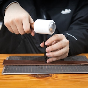 May include: A person using a leather punch and a mallet to make a hole in a piece of brown leather.