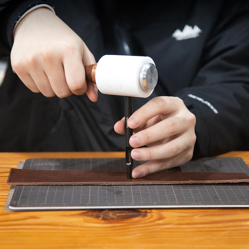 May include: A person using a leather punch and a mallet to make a hole in a piece of brown leather.