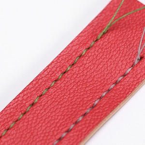 May include: Close-up of a red leather strap with green and gray stitching. The leather has a textured surface. The stitching is visible, showing the craftsmanship of the item. The strap is likely for a watch or other accessory.