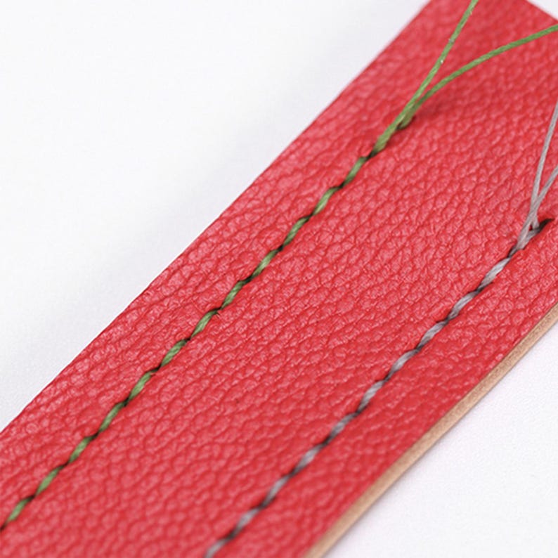 May include: Close-up of a red leather strap with green and gray stitching. The leather has a textured surface. The stitching is visible, showing the craftsmanship of the item. The strap is likely for a watch or other accessory.
