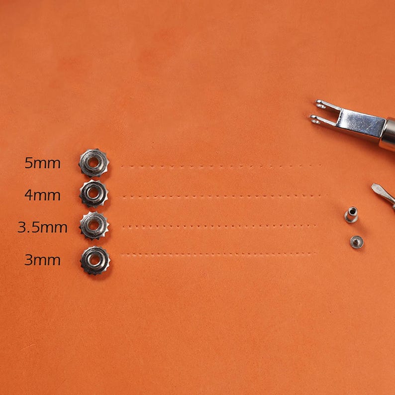 May include: Four silver metal grommets of different sizes, 5mm, 4mm, 3.5mm, and 3mm, are arranged vertically on an orange surface. The grommets are used for leather craft projects.