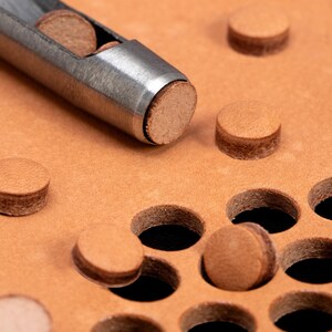 May include: A close-up of a leather punch tool with a metal handle and a round, brown leather punch. The tool is being used to punch holes in a piece of brown leather. The leather has a grid of holes already punched in it.