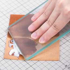 May include: A hand using a clear glass scraper to smooth out a white adhesive on a piece of brown leather. The leather is on a gridded surface.