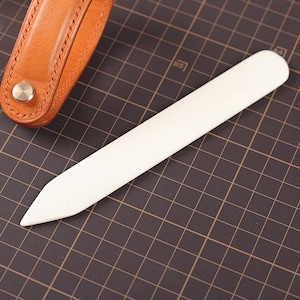 May include: A white leather crafting tool with a pointed tip and rounded end. The tool is lying on a black gridded surface.