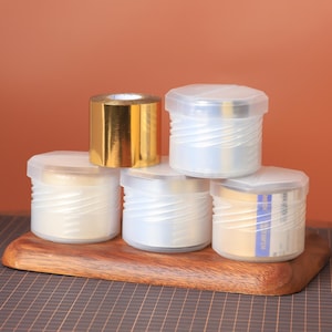 May include: A collection of clear plastic containers with white contents and gold foil tape rolls. The containers have a ridged design and clear lids. The items are arranged on a wooden cutting board against an orange background.