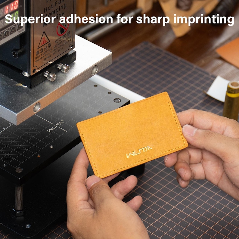 May include: A mustard-yellow leather cardholder with the brand name "WUTA" embossed in gold. The cardholder is being held near a hot foil stamping machine. The text "Superior adhesion for sharp imprinting" is above the cardholder.