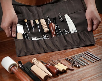 May include: A brown canvas tool roll filled with various leatherworking tools, including knives, awls, and punches. Additional tools are arranged on a wooden surface, such as a mallet, edge beveler, and stitching groover. The tools are for leathercraft.