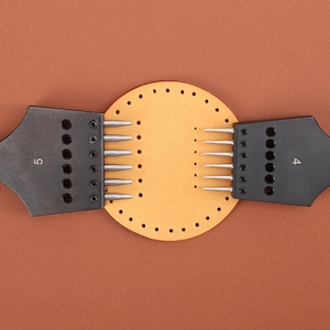 May include: A leather craft tool with two black handles and a circular piece of leather with holes punched around the edge. The handles have rows of metal needles and the numbers "4" and "5" are printed on the handles.