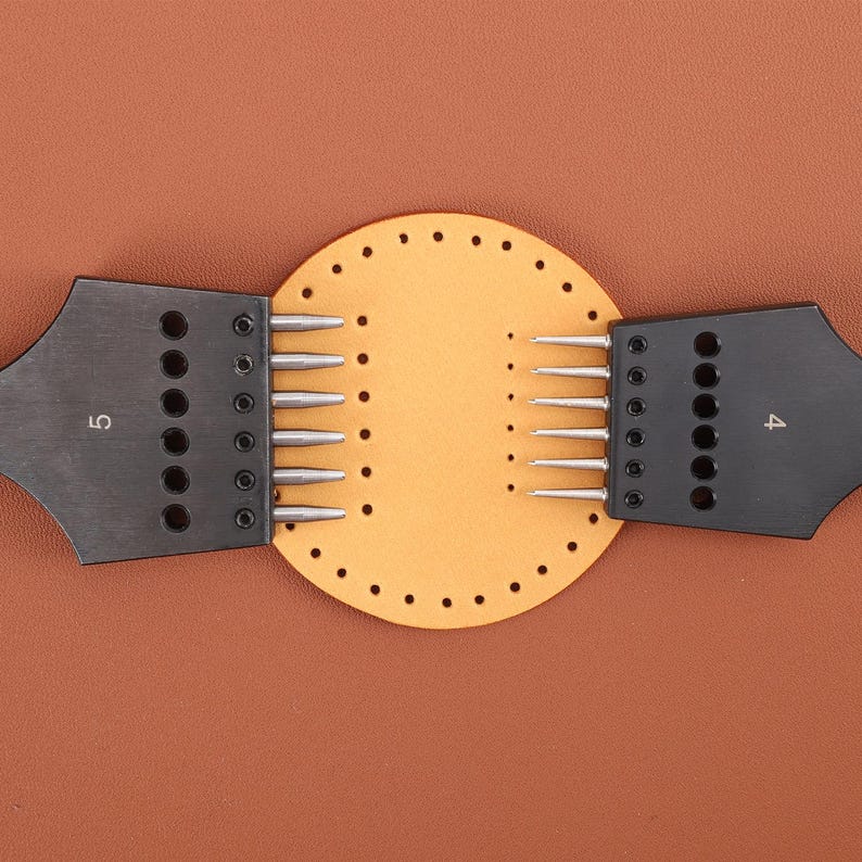 May include: A leather craft tool with two black handles and a circular piece of leather with holes punched around the edge. The handles have rows of metal needles and the numbers "4" and "5" are printed on the handles.