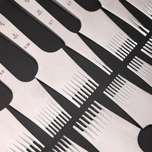 May include: Set of 7 leather stitching tools with different sized needles. The tools are made of metal and have a silver finish. The needles are arranged in a row, with the largest needle on the left and the smallest needle on the right. The needles are labeled with their size in stitches per inch (SPI).