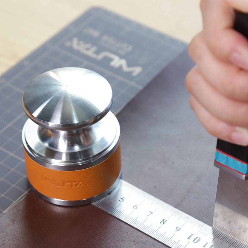 May include: A stainless steel leathercraft tool with a brown leather band and the brand name "WUTA" embossed on it. A metal ruler is placed next to the tool, with a knife in the background. The tool is used for leather cutting and crafting.