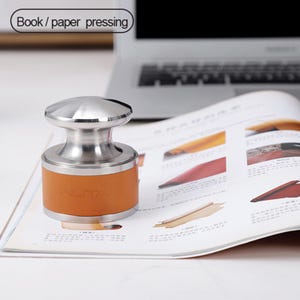 May include: A stainless steel book press with a brown leather band. The press is sitting on a magazine with colorful images. The text "Book / paper pressing" is in the upper left corner.