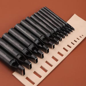 May include: A set of black metal leathercraft punches with various sizes, including 2x6mm, 3x8mm, and 4x10mm, arranged on a tan leather piece with rectangular cutouts. The tools are designed for leatherworking.