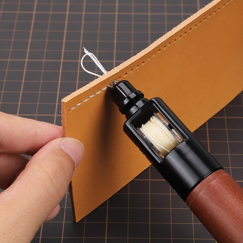 May include: A close-up of a leather sewing awl with a wooden handle and a spool of white thread. The awl is being used to sew a piece of brown leather.
