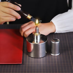 May include: A person uses a small tool to heat a metal container with a flame. The container is sitting on a black and white grid surface. A red piece of leather is in the background.
