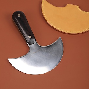 May include: A curved, stainless steel leatherworking knife with a dark wooden handle and brass rivets. The blade is marked with the word "WUTA" and the number "3". A tan leather sheath is in the background.
