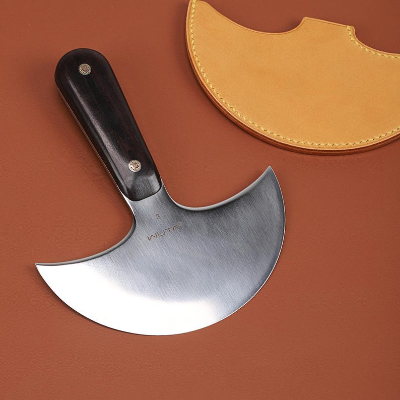May include: A curved, stainless steel leatherworking knife with a dark wooden handle and brass rivets. The blade is marked with the word "WUTA" and the number "3". A tan leather sheath is in the background.