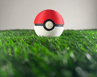 May include: A red and white Poké Ball sits on a bed of green artificial grass. The ball has a black band around the middle with a white button in the center. The top half is red, and the bottom half is white.