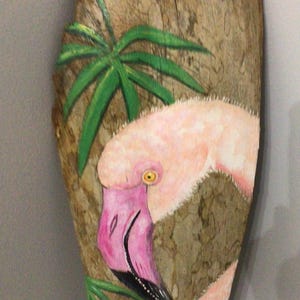 May include: A pink flamingo painted on a piece of wood with green palm leaves in the background.