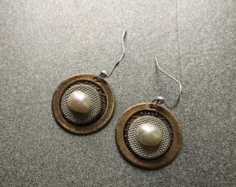 Op de afbeelding: Een paar gouden en zilveren oorbellen met een witte parel in het midden van elke oorbel. De oorbellen hebben een rond ontwerp met een getextureerd oppervlak.