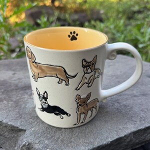 May include: Cream-colored ceramic mug featuring illustrations of various dog breeds. The interior is yellow, with a black paw print at the top. The mug has a white handle and is set against a natural background. A Dachshund, Chihuahua, and French Bulldog are among the breeds depicted.