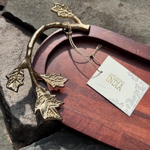 May include: A wooden serving tray with a dark brown finish and gold-colored leaf-shaped handles. The tray has an oval shape and a tag that reads "HANDCRAFTED IN INDIA".