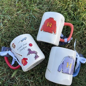 Puede incluir: Tres tazas de cerámica blanca con asas coloridas y gráficos temáticos. Una taza presenta un suéter rojo con una "R" dorada, otra tiene un suéter morado con una "H" dorada, y la tercera tiene varias pegatinas temáticas de magos.