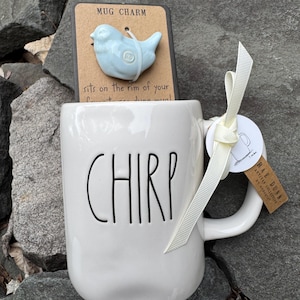 May include: A white ceramic mug with the word "CHIRP" in black lettering. A light blue bird-shaped mug charm is attached to a brown card. A cream-colored ribbon and tag are tied to the mug's handle.
