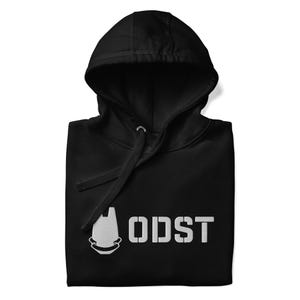 May include: Black hooded sweatshirt with white text and a graphic. The text reads "ODST" in a bold, sans-serif font. The graphic is a stylized helmet design. The hoodie has a drawstring and a front pocket.