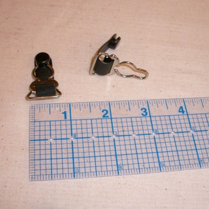 May include: Two black suspender clips with silver-colored metal accents. One clip is closed, and the other is open. A blue ruler with inch markings is in the foreground, showing measurements.