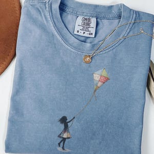 Puede incluir: Una camiseta azul Comfort Colors con la silueta de una niña volando una cometa. La cometa es multicolor con cuadrados rojos, verdes y amarillos. Un collar dorado con un colgante está drapeado sobre la camisa. Un sombrero marrón es visible en el fondo.