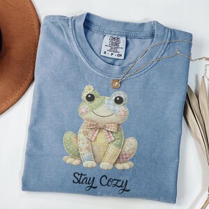 Può includere: Una maglietta Comfort Colors azzurra con una grafica di rana patchwork, un papillon e una margherita. Il testo "Stay Cozy" è stampato sotto la rana. Una collana d'oro con un ciondolo è drappeggiata sulla maglietta.