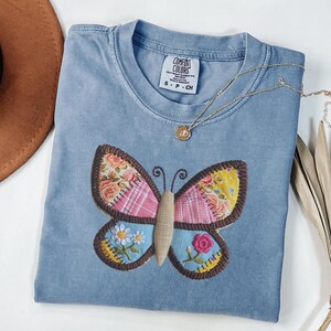 Könnte beinhalten: Hellblaues Comfort Colors T-Shirt mit einem bunten Schmetterlingsaufnäher. Der Schmetterling hat rosa, gelbe und blaue Bereiche mit Blumen- und Karomustern. Eine goldene Halskette mit Anhänger liegt auf dem Shirt.