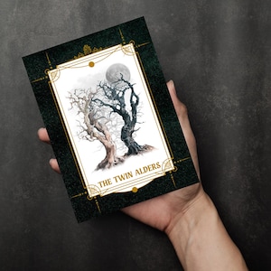 Pode incluir: Um cart&atilde;o verde escuro com uma borda dourada e o texto "THE TWIN ALDERS". O cart&atilde;o apresenta uma ilustra&ccedil;&atilde;o de duas &aacute;rvores, uma clara e outra escura, sob uma lua cheia. O cart&atilde;o &eacute; segurado em uma m&atilde;o.