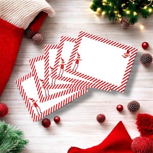 May include: A stack of white rectangular cards with red and white diagonal stripes and a small elf illustration. The cards are arranged on a light wood surface, surrounded by red baubles, a red stocking, and Christmas greenery.